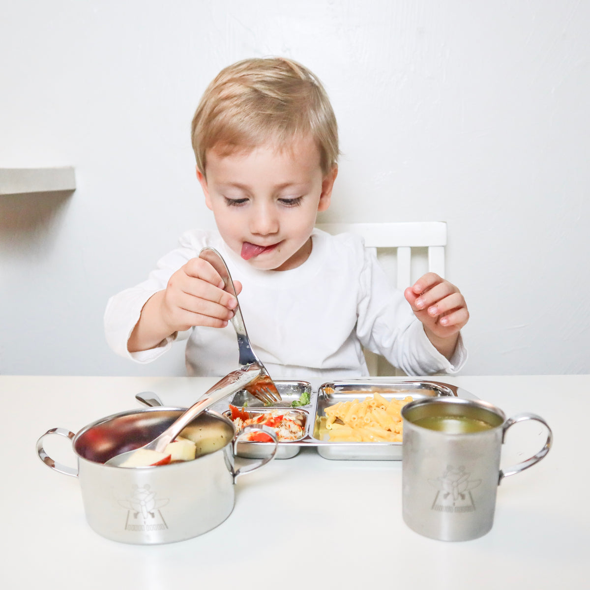 A toddler boy is eating from a set of stainless steel dinnerware consists of a stainless steel divided tray, a stainless steel bowl with handles, and a stainless steel cup with handle.