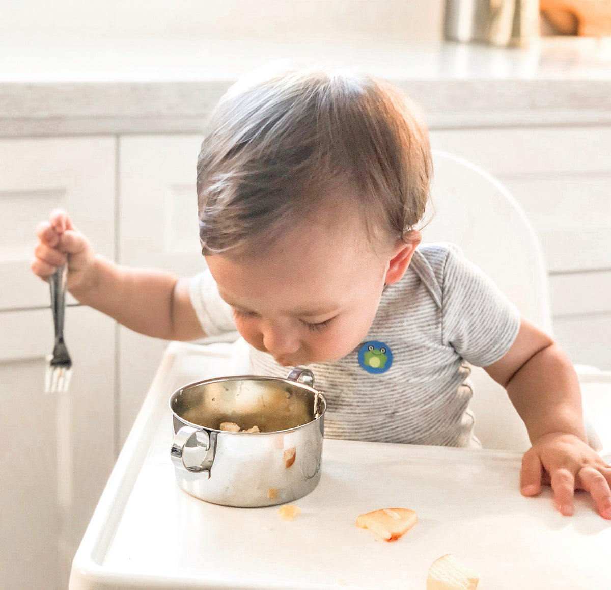 A baby is eating food from a Kiddobloom baby safe stainless steel bowl with two handles and a baby safe stainless steel fork. The bowl and fork have smooth edges and luxurious mirror polish.